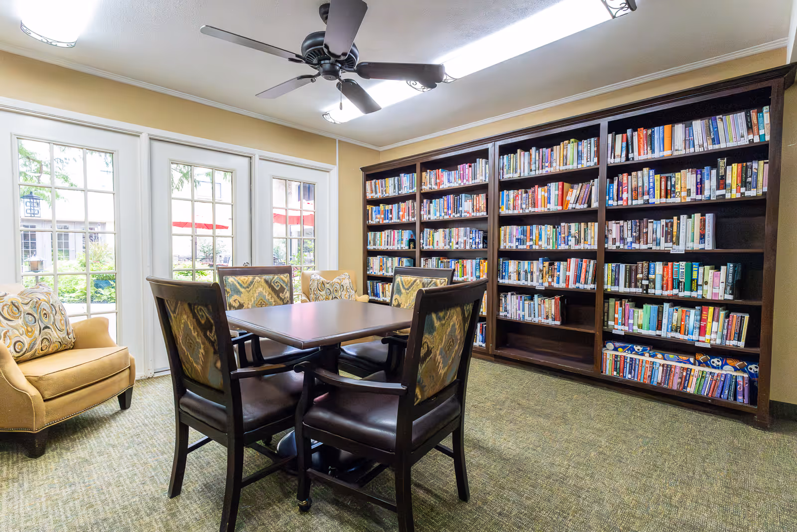 A cozy reading room with a large bookshelf filled with books along one wall, a square wooden table surrounded by four cushioned chairs with patterned backs, a beige armchair with a decorative pillow, and large glass doors letting in natural light and showing an outdoor garden area.