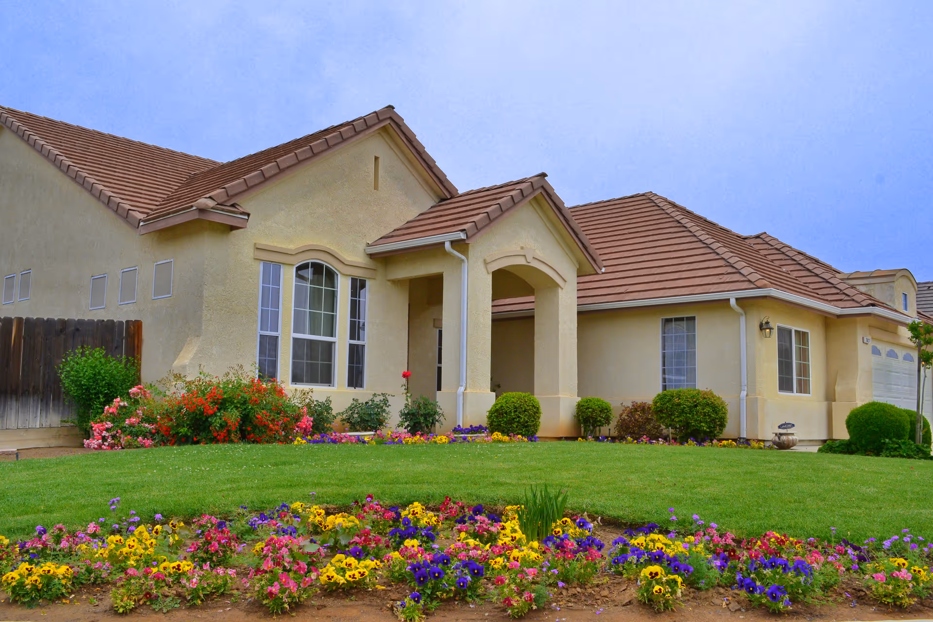 Single-story residential care facility with beige stucco walls and a brown tiled roof, surrounded by a well-maintained lawn and colorful flower beds in front under a clear blue sky.