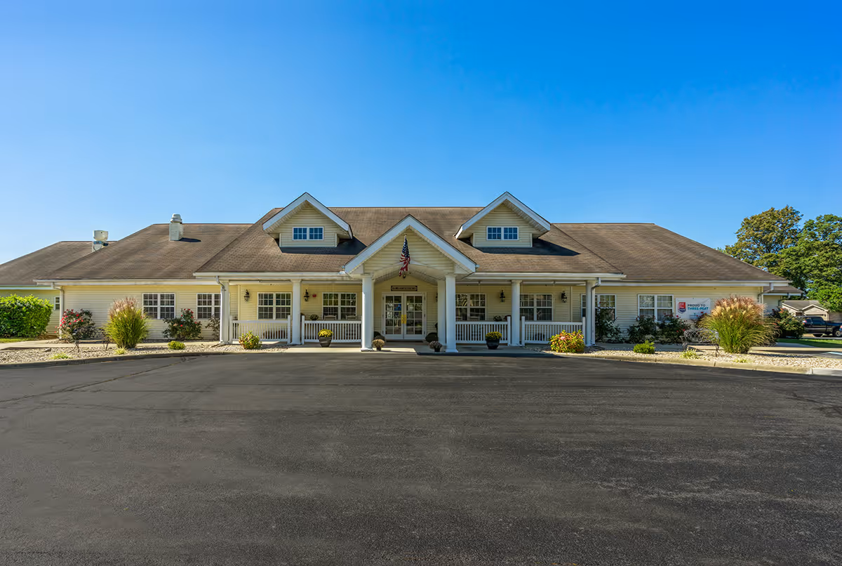 Front exterior view of a single-story building with a beige facade, a brown shingled roof, and a covered entrance supported by white columns. The building is surrounded by small shrubs and plants, with a clear blue sky overhead.