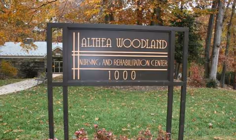 A black sign reading Althea Woodland Nursing and Rehabilitation Center 1000 on a grassy lawn.