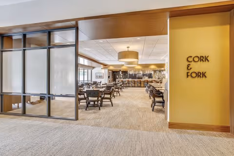 Entrance to a dining area named Cork & Fork in Greenspring Senior Living Community, featuring multiple tables and chairs arranged neatly with a modern and spacious interior design.