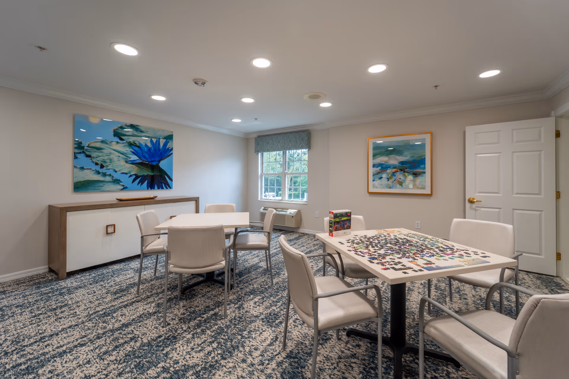 A well-lit activity room with two tables surrounded by beige chairs. One table has a partially completed jigsaw puzzle and a puzzle box on it. The room features patterned carpet, two framed artworks on the walls, a window with a blue valance, and a white door with a gold handle.