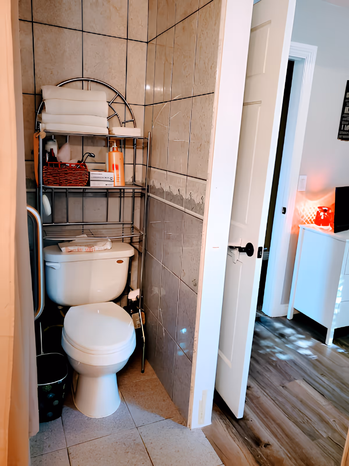 A small bathroom with a white toilet and a metal shelf above it holding folded towels, a basket, and various toiletries. The walls are tiled, and the bathroom door is open, showing a glimpse of a room with wooden flooring and a white dresser with a red lamp on top.