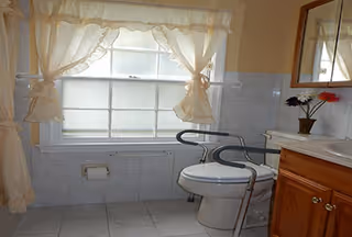 Bathroom with a toilet fitted with supportive grab bars, a wooden vanity with mirror, tiled walls and floor, and a curtained window.