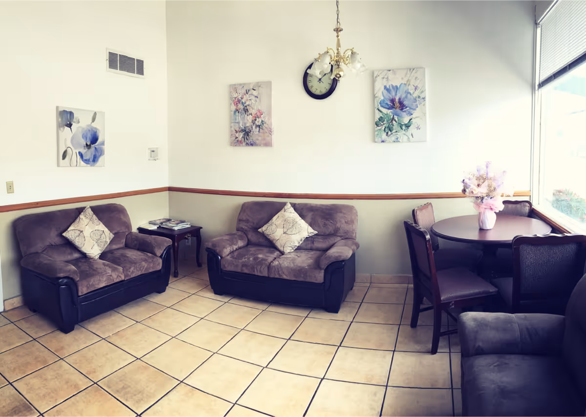 A cozy sitting area with two brown cushioned loveseats, each with a decorative pillow, a small wooden side table with magazines, a round wooden table with four chairs, and a vase with flowers. The walls are decorated with three floral paintings and a clock. The floor is tiled, and there is a large window letting in natural light.