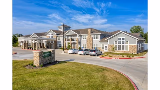 Exterior view of Cedarhurst Senior Living of Blue Springs building with a stone and siding facade, several parked cars, a driveway, and a green lawn under a blue sky with some clouds.