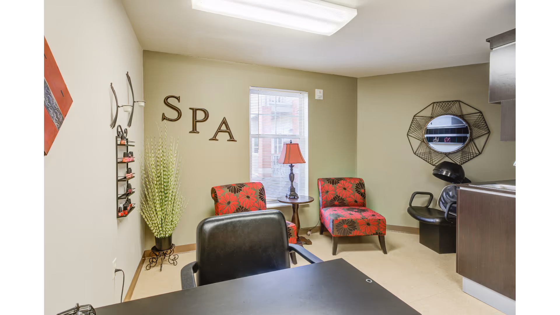 Interior of a spa room with two red and brown floral chairs, a small round table with a red lamp, a black salon chair, a hair washing station with a black chair and sink, a decorative mirror on the wall, a tall plant in a black pot, and the word 'SPA' displayed on the wall.