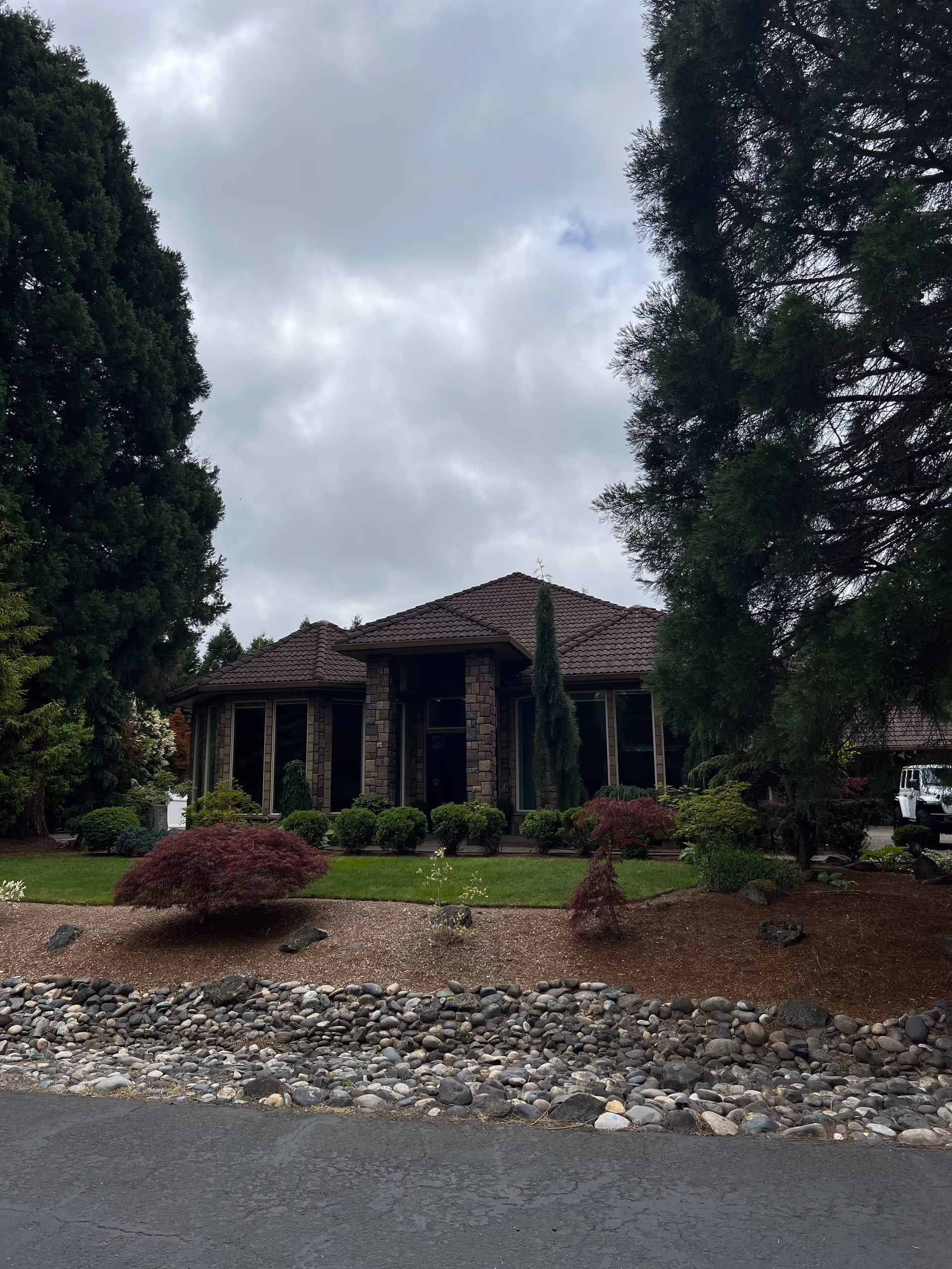 Front exterior of a single-story stone-faced house with manicured landscaping, trees, and a cloudy sky.