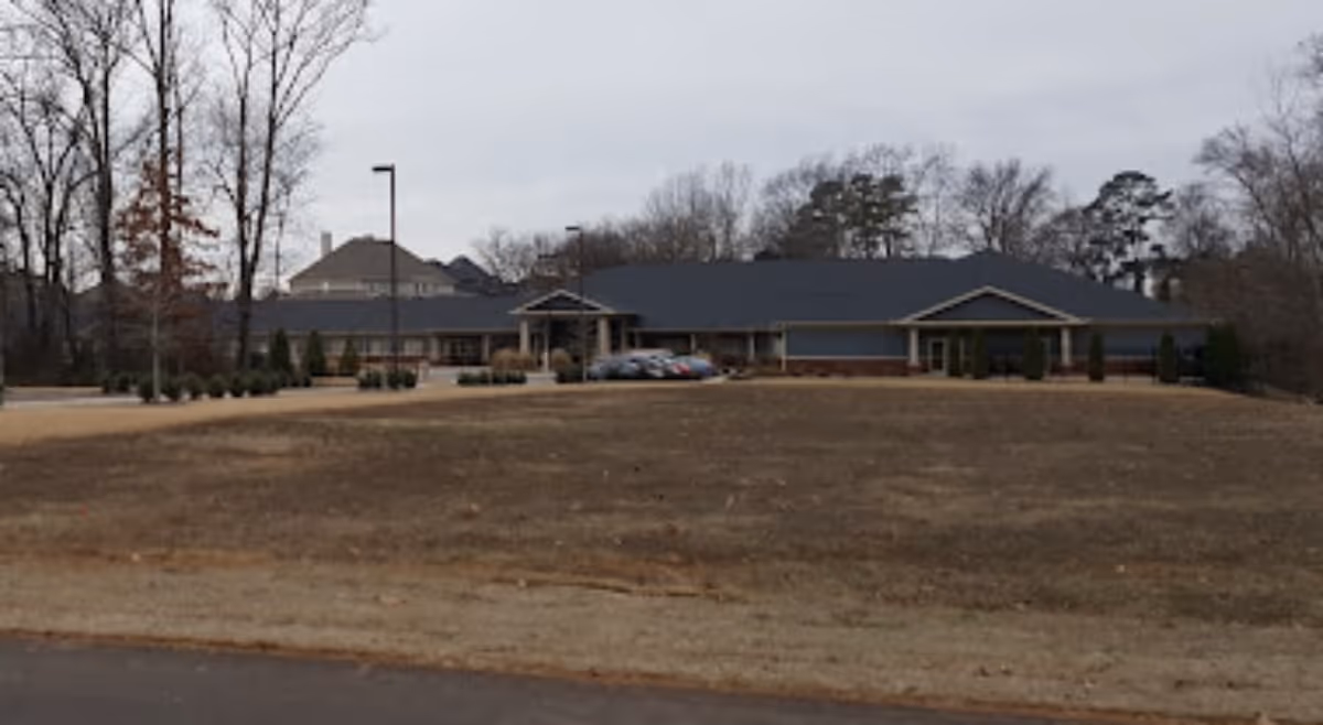 Low single-story assisted living building seen from across a large grassy front lawn with a few parked cars and trees under an overcast sky.