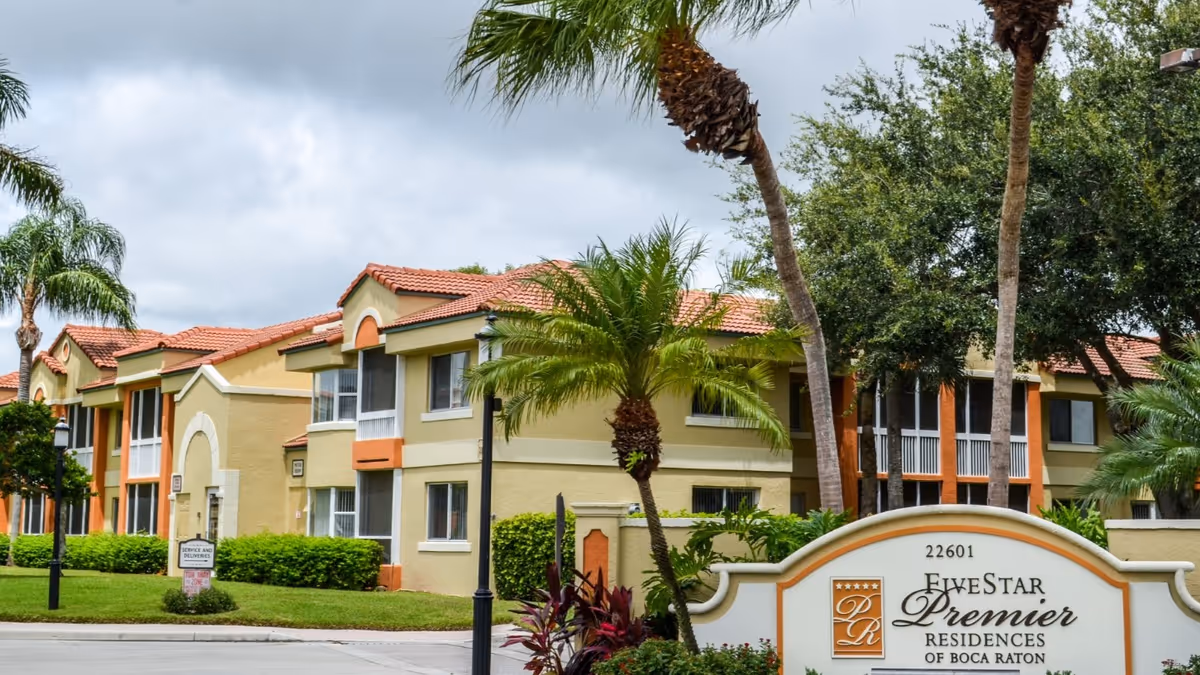 Exterior view of Five Star Premier Residences of Boca Raton showing the entrance sign, palm trees, and Mediterranean-style apartment buildings.