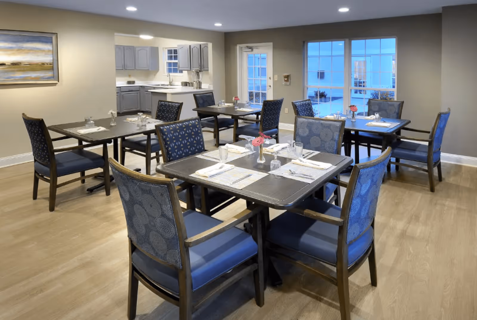 A dining room with four square tables, each set with placemats, napkins, glasses, and small flower vases. The room has wooden flooring, beige walls, and large windows letting in natural light. In the background, there is a small kitchen area with gray cabinets and a countertop.