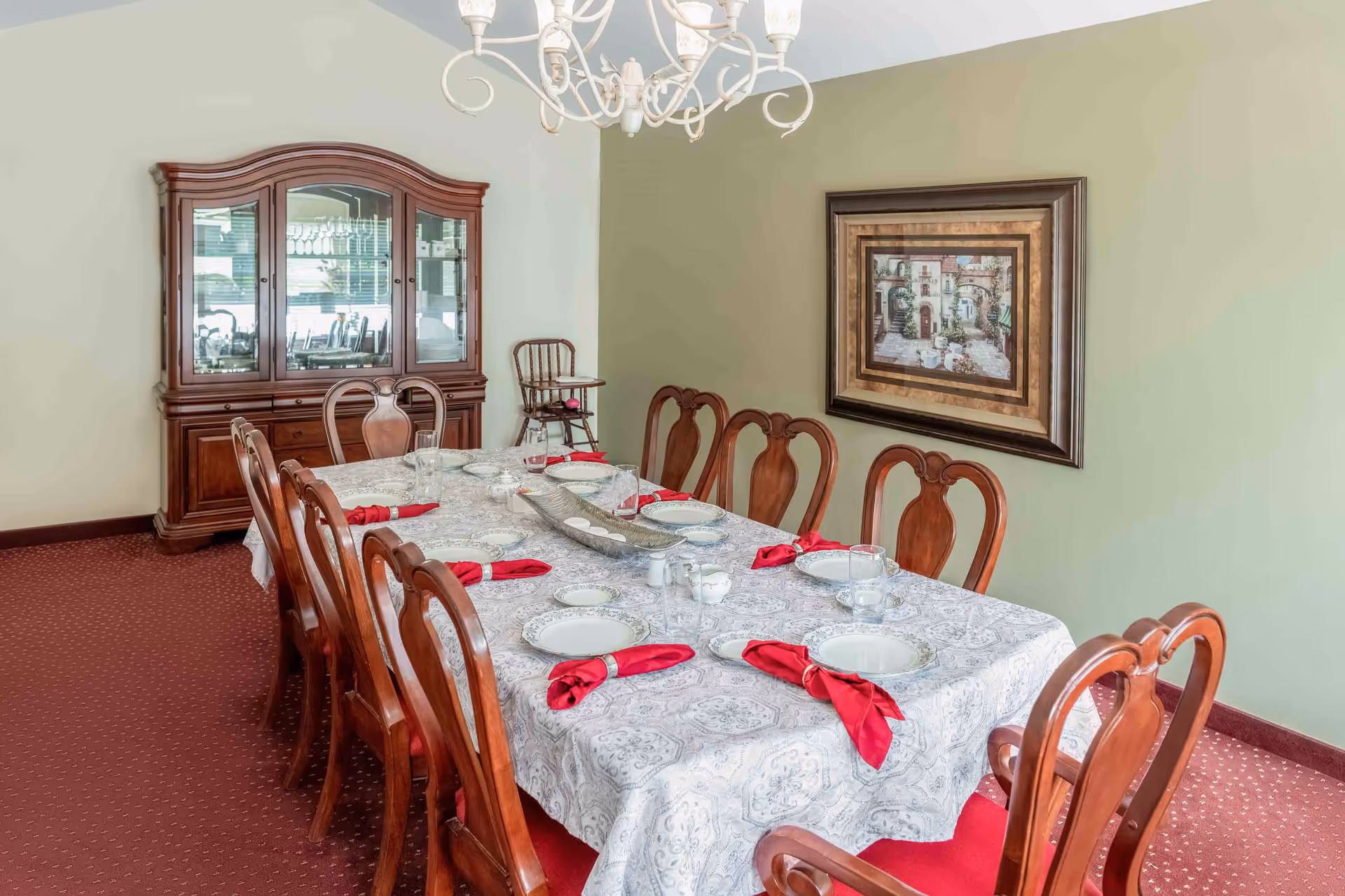 A formal dining room with a long table covered by a patterned tablecloth, set with plates, glasses, and red napkins. There are eight wooden chairs around the table, a wooden china cabinet in the corner, a high chair against the wall, and a framed painting on a light green wall. A chandelier hangs above the table.