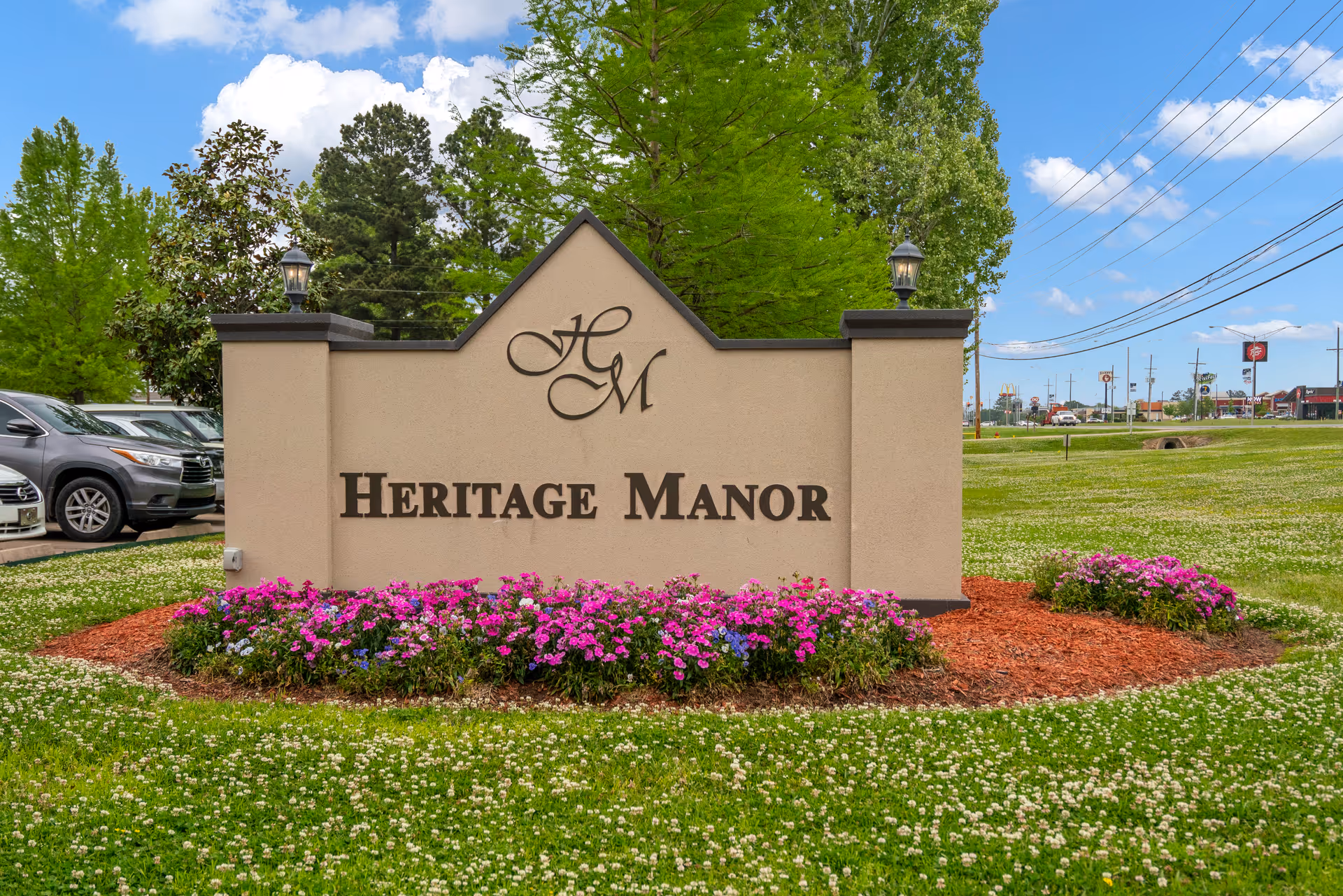 A large beige stone sign with the letters 'HM' and the words 'Heritage Manor' in dark brown, surrounded by green grass and colorful flowers, with trees and a partly cloudy sky in the background.