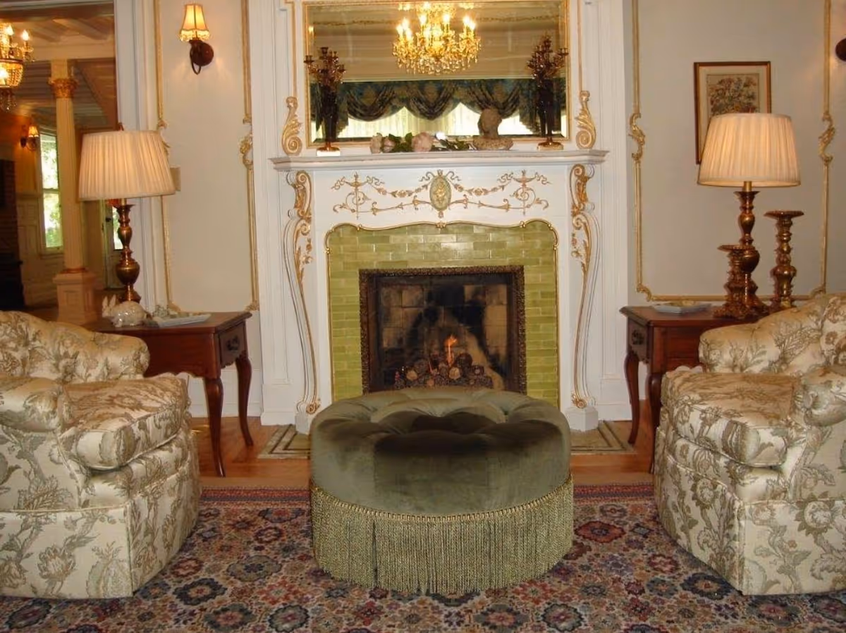 A cozy living room with a decorative fireplace featuring green tiles and ornate white and gold detailing. In front of the fireplace is a round green upholstered ottoman with fringe. On either side of the fireplace are floral patterned armchairs and wooden side tables with brass lamps. A large mirror hangs above the fireplace reflecting a chandelier and window with drapes.