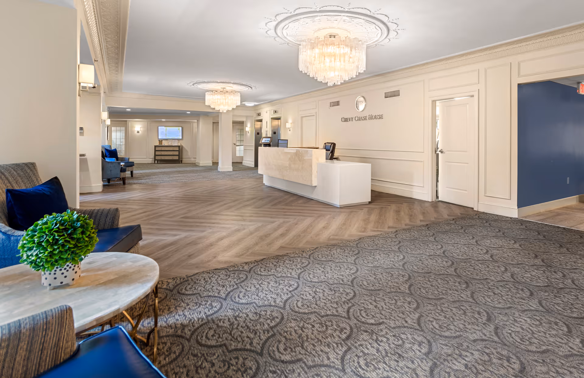 Bright, elegant reception lobby with a marble front desk, crystal chandeliers, seating area, and patterned carpeting.