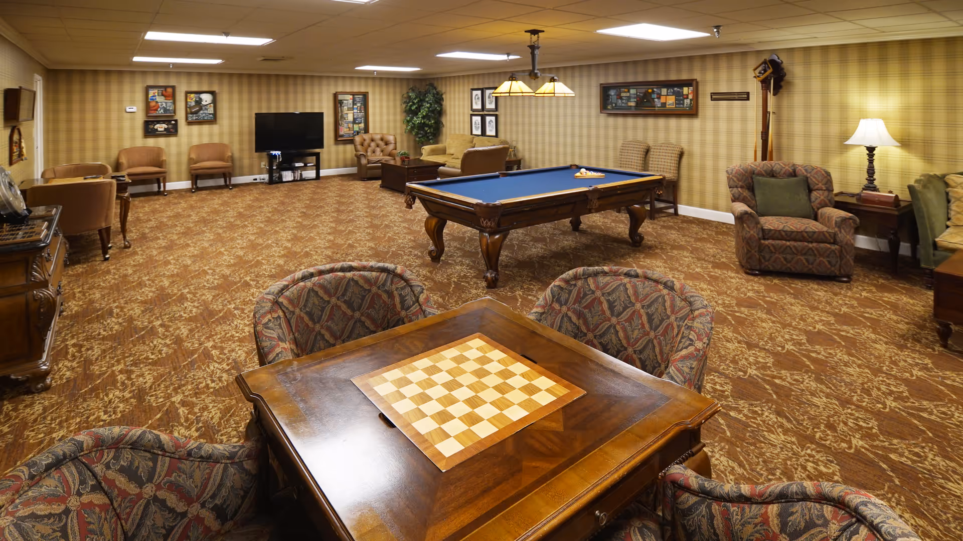 A spacious senior living community game room with a pool table in the center, a wooden table with a chessboard in the foreground, several upholstered chairs and sofas arranged around the room, a flat-screen TV mounted on the far wall, framed pictures and plants decorating the walls, and warm lighting from ceiling fixtures and a table lamp.
