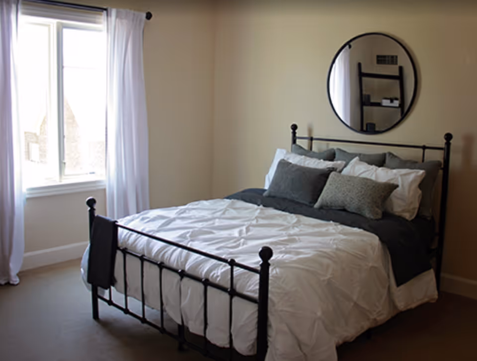 A bedroom with a black metal bed frame, white comforter, and multiple pillows in shades of gray and white. There is a round mirror on the wall above the bed and a window with white curtains letting in natural light.