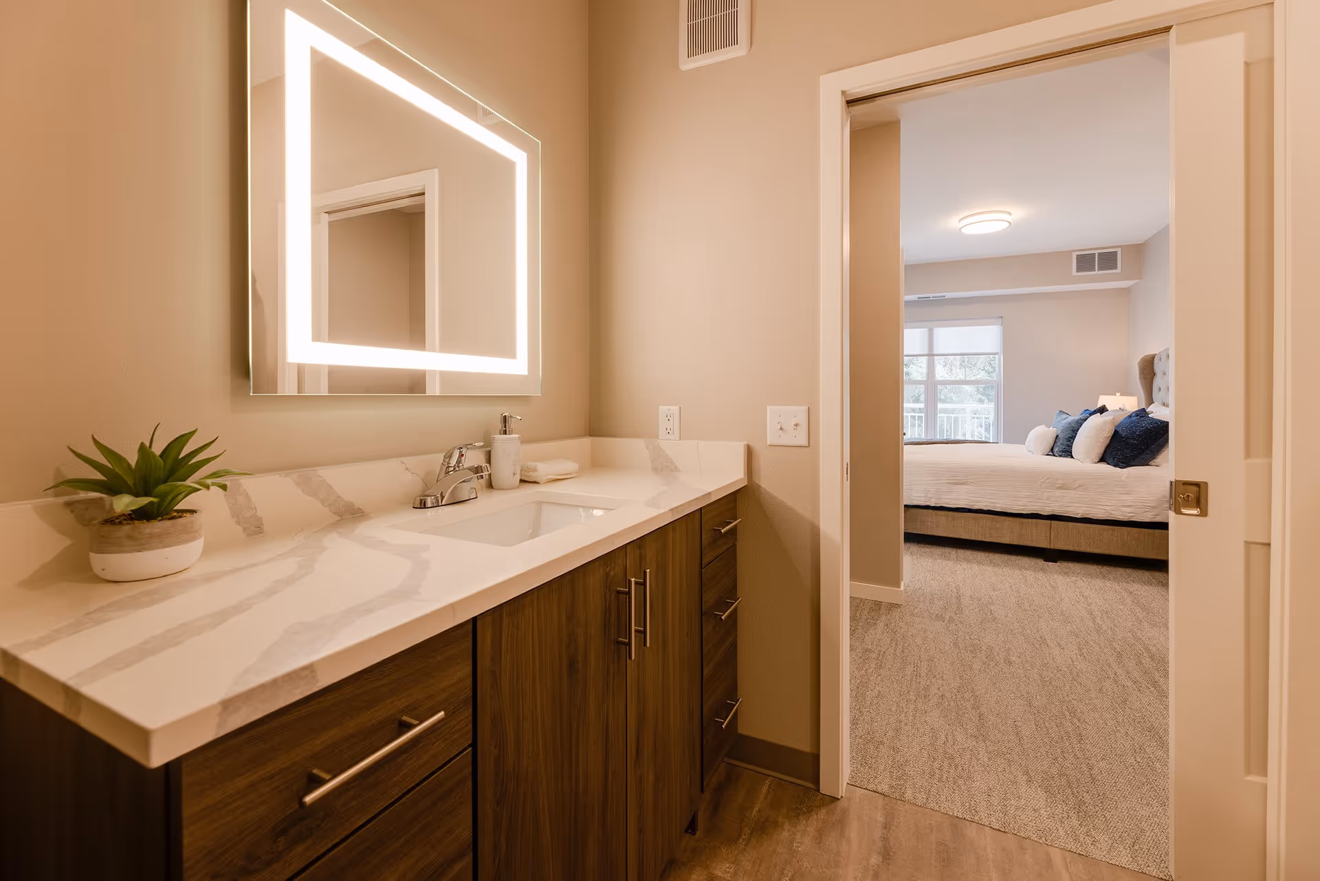 A modern bathroom vanity with a white marble countertop, a built-in sink, and a large illuminated mirror above. A small potted plant and a soap dispenser are on the countertop. The bathroom opens into a bedroom with a bed, pillows, a window, and carpeted flooring.