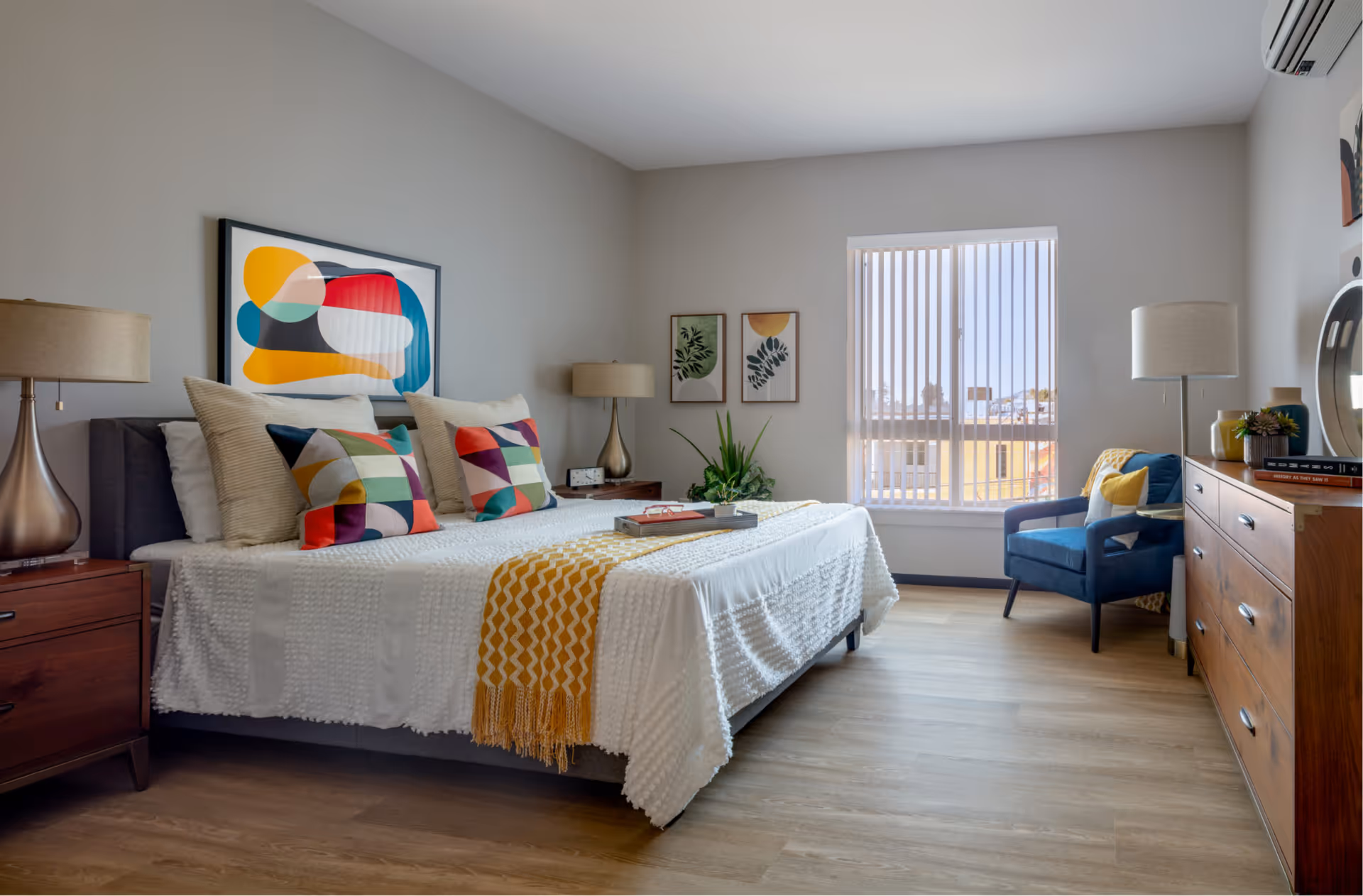 A bright and modern bedroom with a large bed adorned with white bedding, colorful geometric pillows, and a yellow throw blanket. The room features wooden nightstands with lamps on either side of the bed, abstract artwork above the headboard, two framed botanical prints on the wall, a blue armchair with cushions near a window with vertical blinds, and a wooden dresser with decorative items and a round mirror. The floor is light wood, and there is an air conditioning unit mounted on the wall.