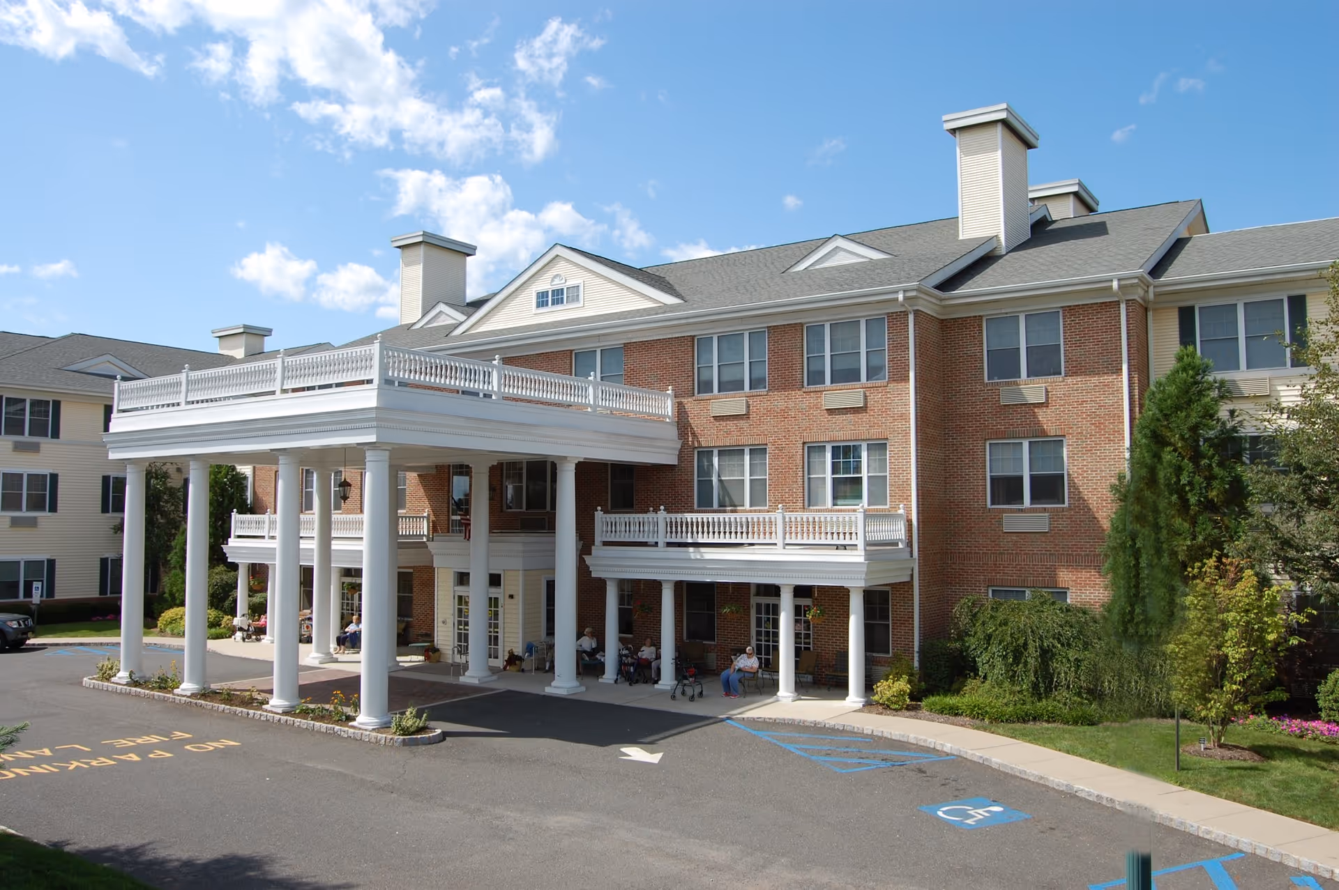 Exterior view of a multi-story assisted living facility with a large covered entrance supported by white columns. There are several people sitting under the covered area near the entrance. The building has a brick facade with multiple windows and a well-maintained surrounding landscape including trees and shrubs. The sky is clear with a few clouds.