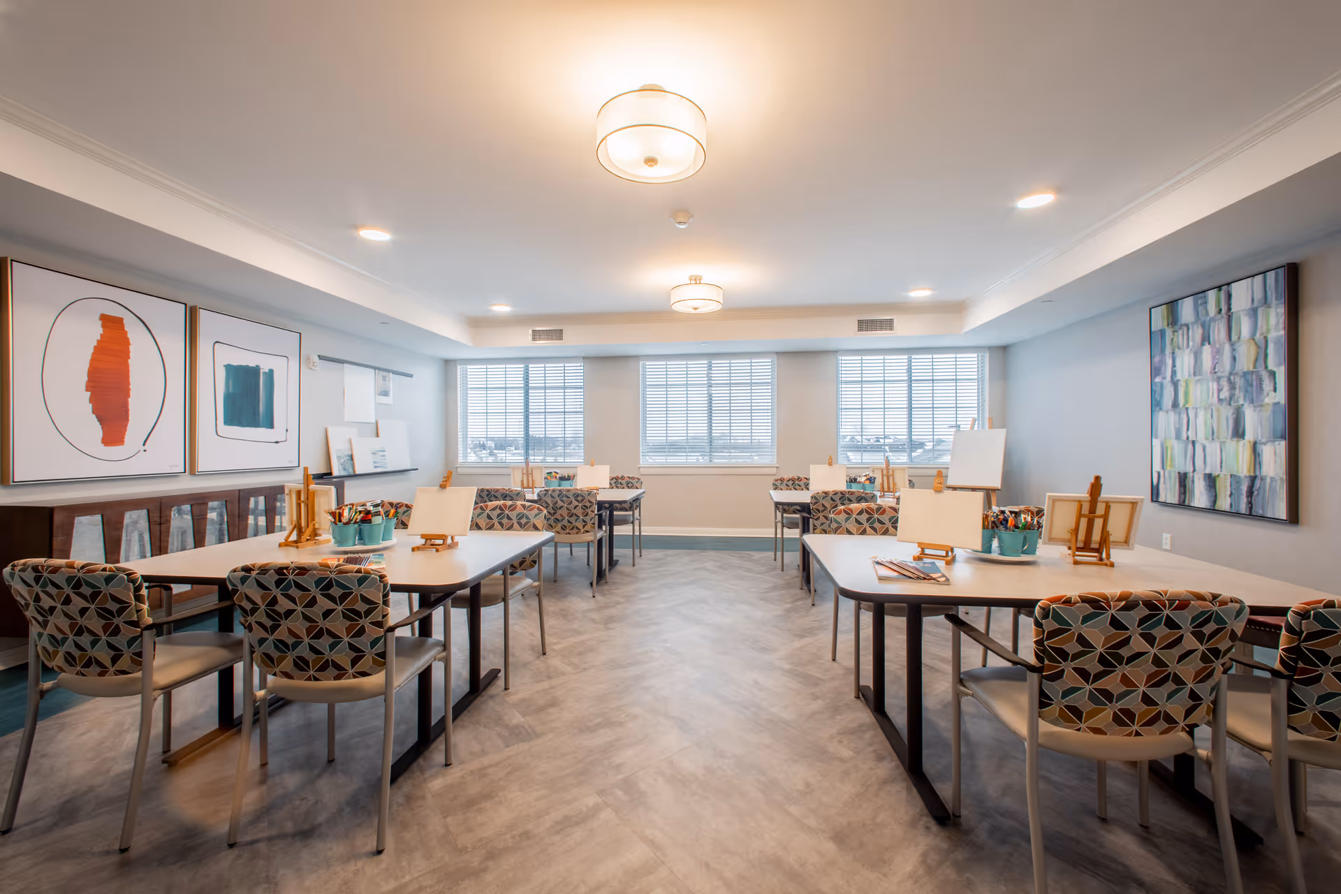 A bright and spacious activity room in a senior living facility with several tables and patterned chairs arranged neatly. Each table has art supplies including paintbrushes, easels, and canvases. The room has large windows with blinds allowing natural light to fill the space. Modern ceiling lights illuminate the room, and abstract artwork decorates the walls.