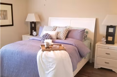 A neatly made bed with a white headboard, covered with a purple quilt and multiple decorative pillows. A wooden tray with a coffee mug and a book is placed on the bed, along with a white throw blanket draped over the edge. Two matching nightstands with modern black lamps and decorative items flank the bed. A framed picture hangs on the beige wall to the left.