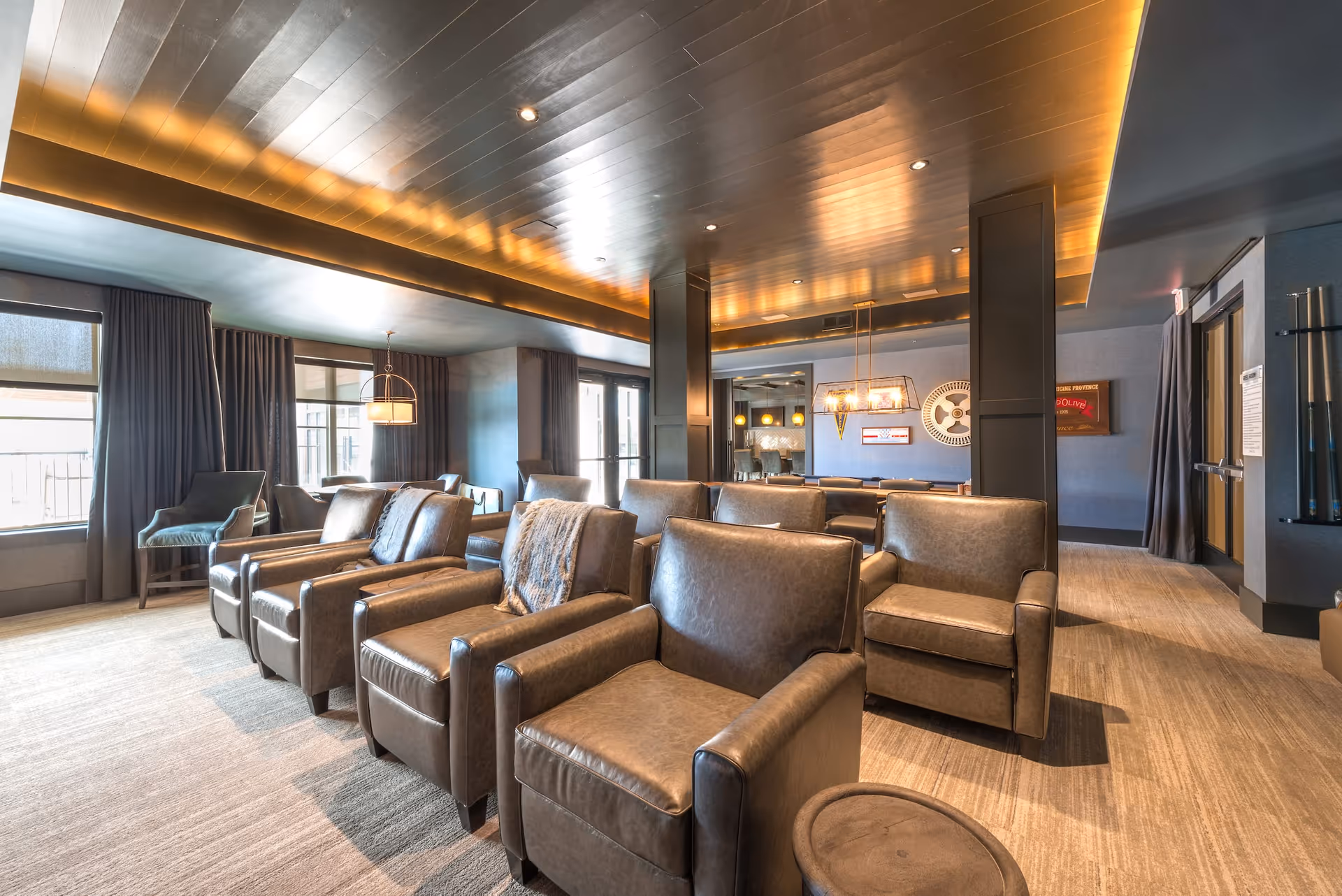 A modern lounge area with multiple brown leather armchairs arranged in rows facing a large window. The room features a wooden ceiling with recessed lighting, large windows with dark curtains, and a carpeted floor. In the background, there is a dining table with chairs and decorative wall art.
