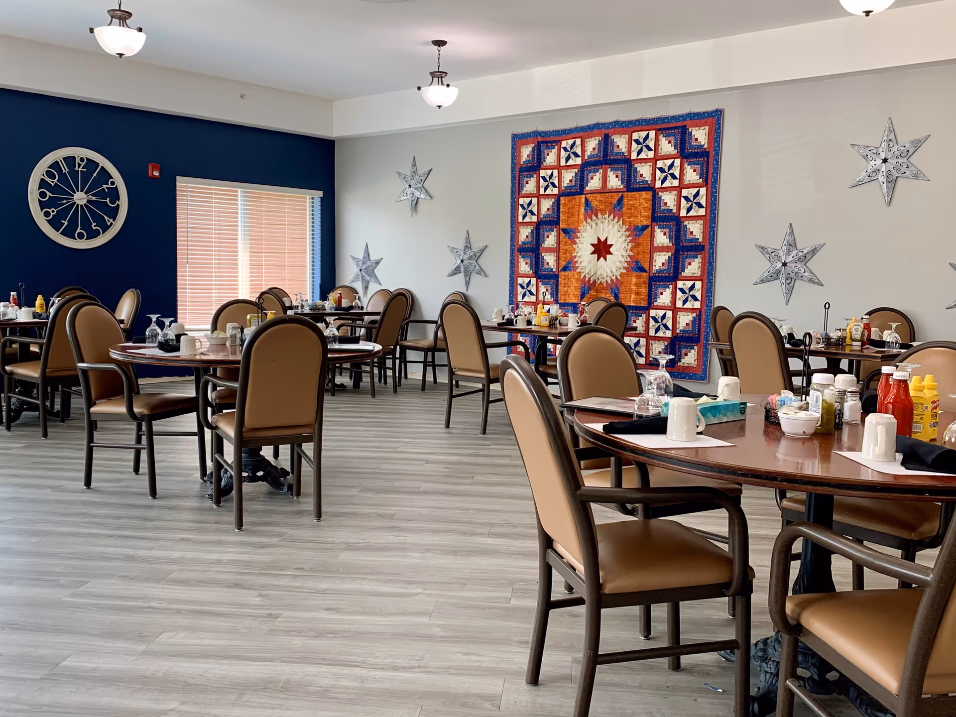 A dining room with several round tables and brown cushioned chairs. Each table is set with cups, napkins, condiments, and glassware. The walls are decorated with a large colorful quilt and several metal star-shaped ornaments. A large clock is mounted on a dark blue accent wall near a window with closed blinds.
