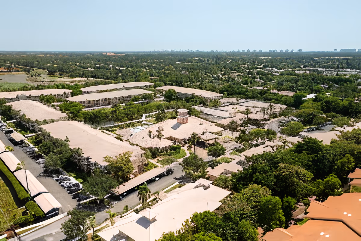 Aerial view of Aston Gardens At Pelican Marsh senior living facility showing multiple low-rise buildings with beige roofs surrounded by green trees and landscaping under a clear blue sky.