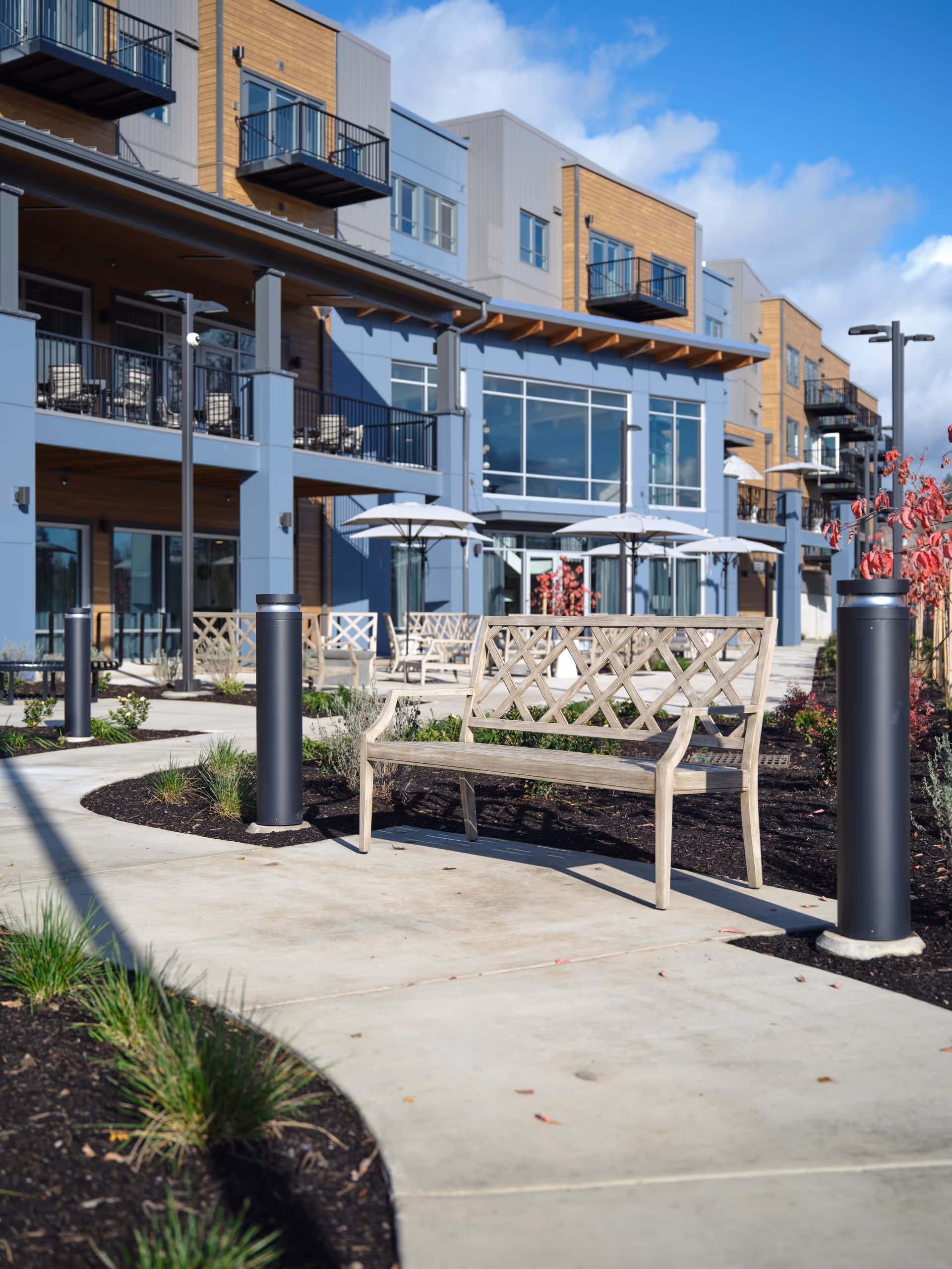 Outdoor patio area of a senior living facility with wooden benches, umbrellas, and landscaped garden beds along a concrete walkway. The building has multiple balconies and large windows under a partly cloudy sky.