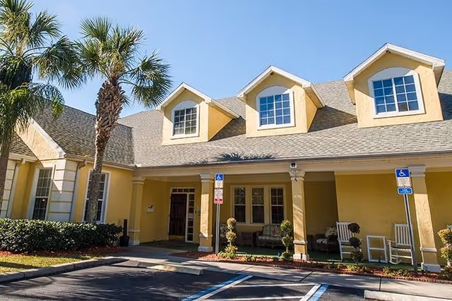 Exterior view of a yellow senior living facility building with three dormer windows on the roof, palm trees, and a covered entrance with rocking chairs and a bench. Handicap parking spaces are visible in front of the building.