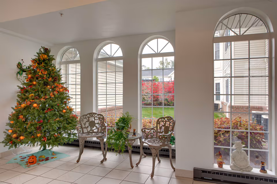 A bright indoor sitting area with four tall arched windows showing an outdoor garden. Inside, there is a decorated Christmas tree with orange lights and fall-themed ornaments, two ornate metal chairs with a matching small table holding potted plants, and a white statue of a seated figure near the window.