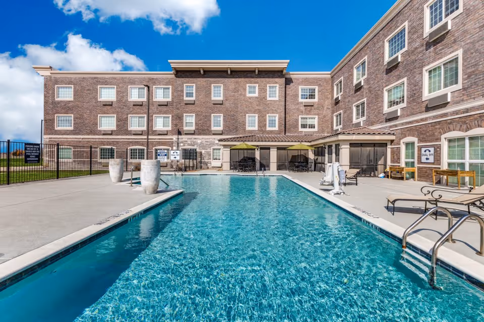 Outdoor swimming pool with clear blue water surrounded by a concrete deck. There are lounge chairs, tables with umbrellas, and a three-story brick building in the background under a partly cloudy blue sky.