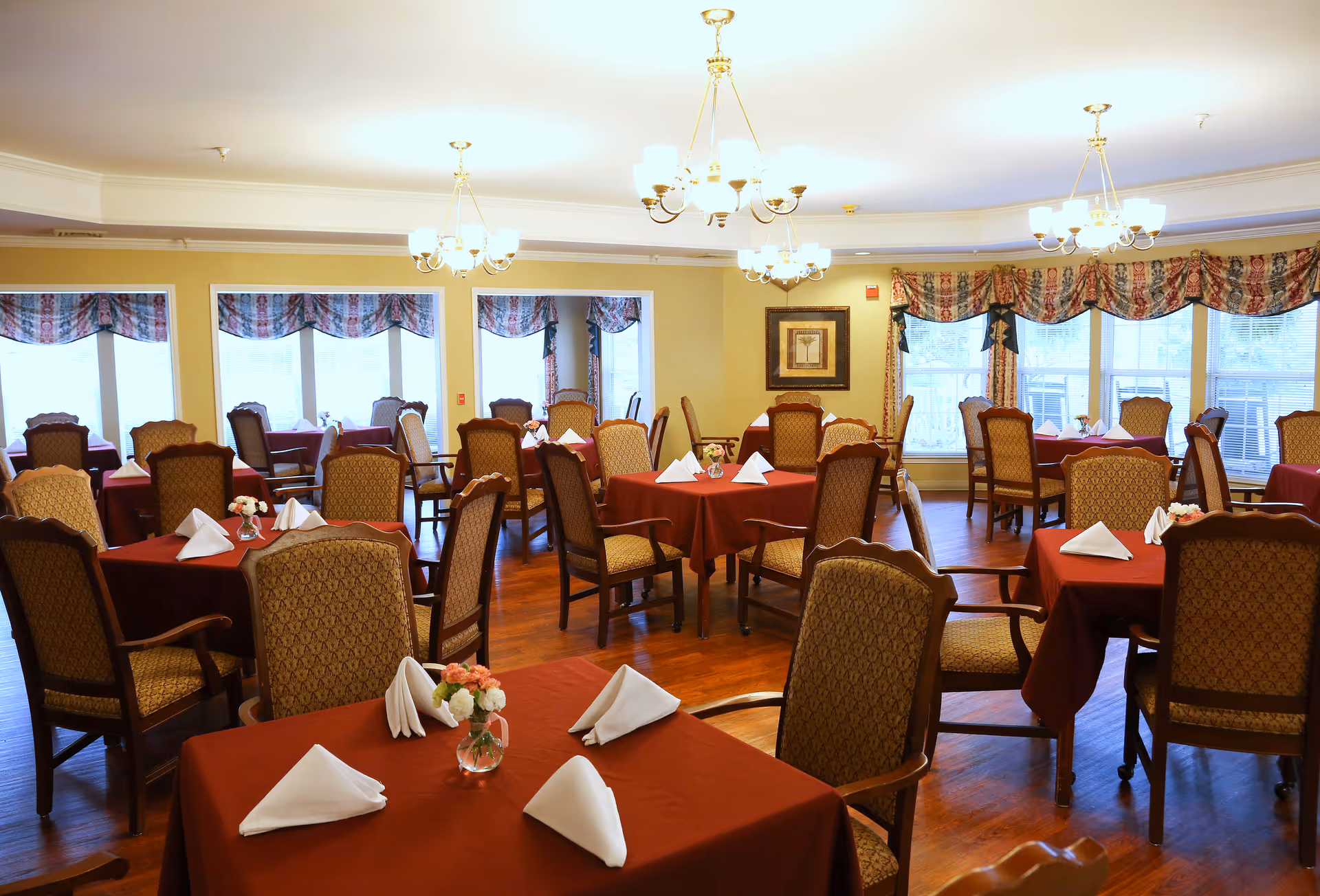 A dining room with multiple tables covered in red tablecloths, each set with white folded napkins and small floral centerpieces. The room has wooden floors, upholstered chairs, large windows with patterned valances, and several chandeliers providing warm lighting.