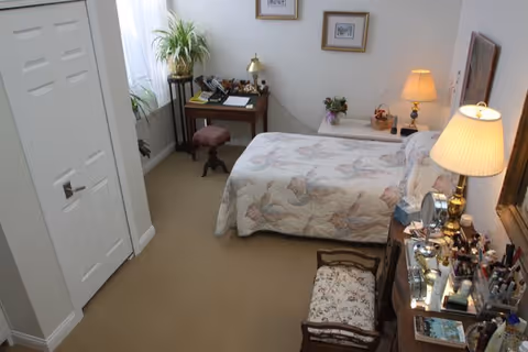 A cozy bedroom with a bed covered in a floral quilt, a bedside table with a lamp and flowers, a small desk with a stool and a plant, framed pictures on the wall, and a dresser with various personal items and a mirror.