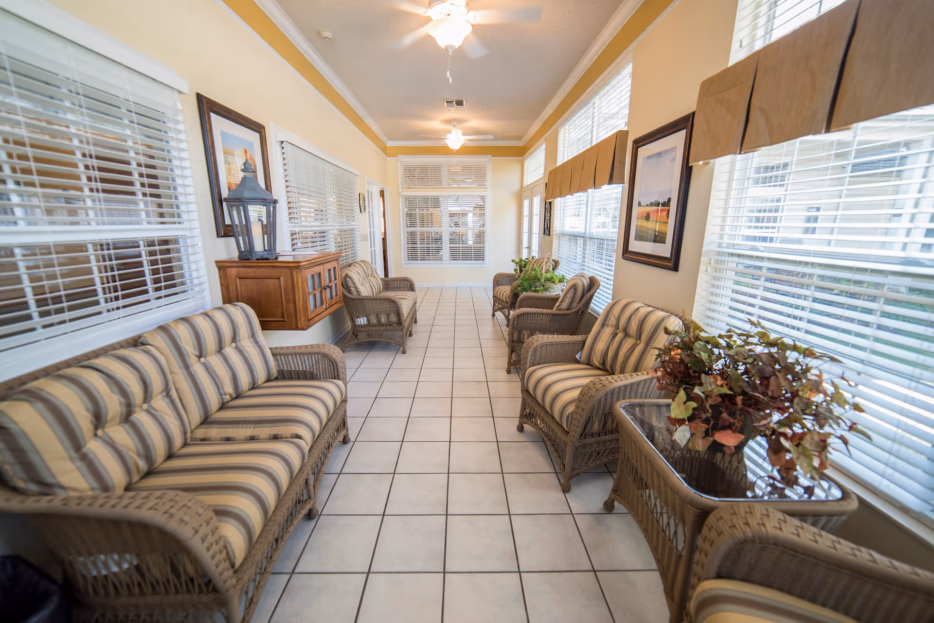 A bright and airy sunroom with large windows on both sides, furnished with wicker sofas and armchairs with striped cushions. There are framed pictures on the walls, a small wooden cabinet with a lantern on top, and potted plants on a glass-top wicker table. The floor is tiled, and ceiling fans with lights are mounted on the ceiling.
