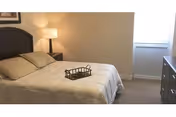 Neatly made bed with pillows and a tray, a bedside table with lamp, dresser, and a window in a simple bedroom.
