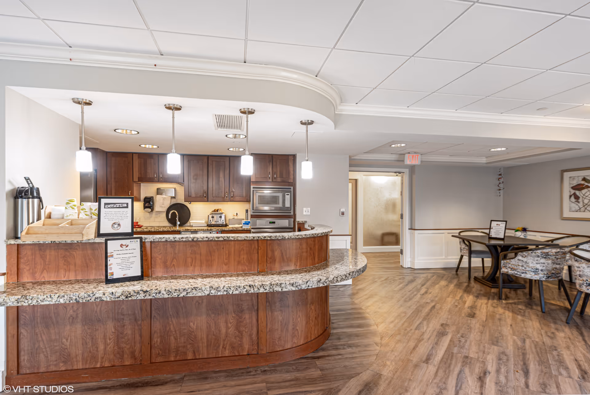 Interior view of a senior living facility's dining and kitchen area featuring a curved granite countertop with wooden cabinetry, three pendant lights hanging above, and a coffee station on the left. In the background, there are tables and chairs for dining, a framed artwork on the wall, and an exit door.