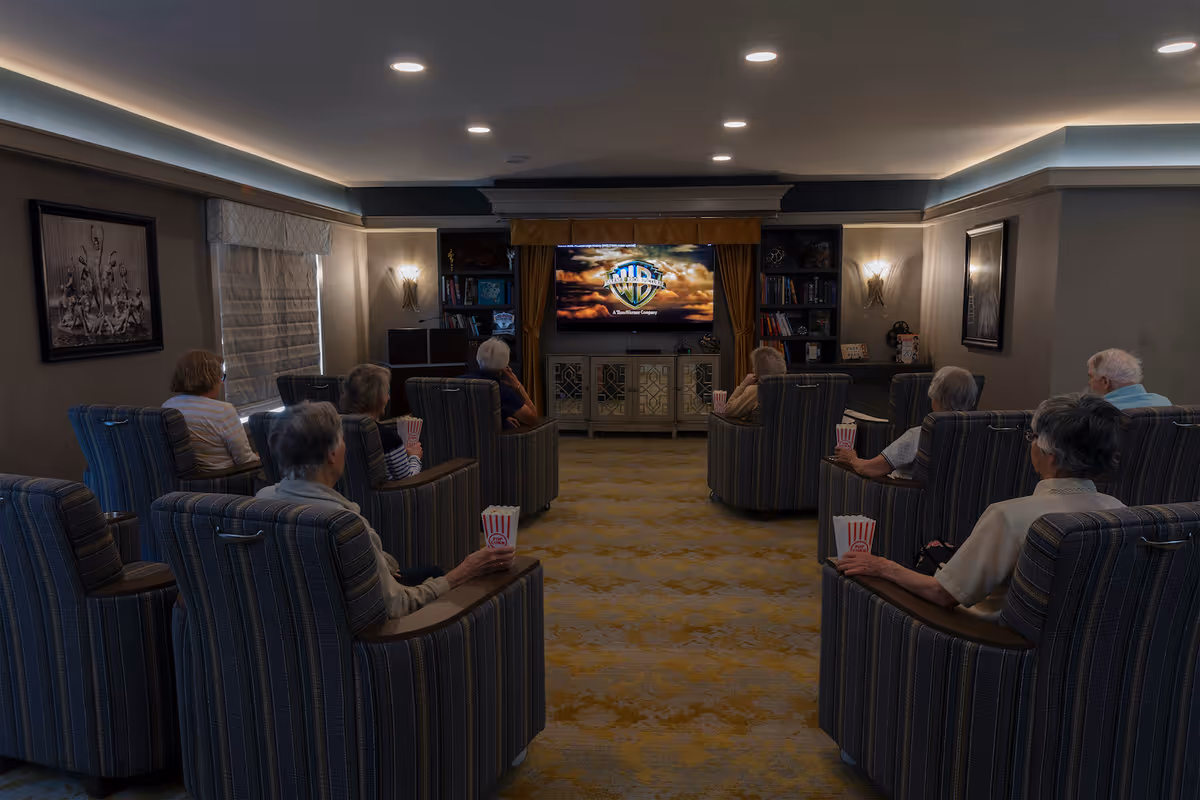 A group of elderly people seated in comfortable armchairs in a dimly lit room, watching a movie on a large flat-screen TV. Each person is holding a container of popcorn. The room has carpeted flooring, bookshelves, and framed artwork on the walls.