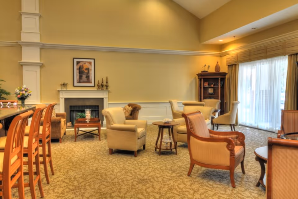 A cozy living room area in a senior living facility with beige armchairs and wooden chairs arranged around small round tables. The room features a fireplace with a framed picture above it, a tall wooden cabinet in the corner, and large windows with sheer curtains allowing natural light to fill the space. The carpet has a subtle patterned design, and the walls are painted a warm yellow color.