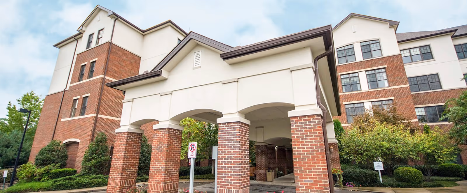 Exterior view of a multi-story senior living facility building with a covered entrance supported by brick columns. The building features a combination of red brick and light-colored siding, surrounded by landscaped greenery and trees under a partly cloudy sky.