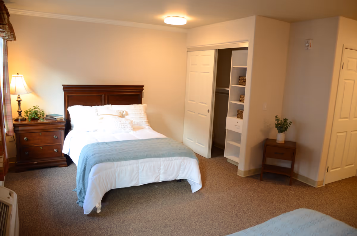 A cozy bedroom with a neatly made bed featuring white bedding and a light blue throw blanket. There is a wooden headboard and a matching wooden nightstand with a lamp, a plant, and some books on it. The room has beige walls, carpeted floor, a closet with sliding white doors, and a small wooden side table with a potted plant.