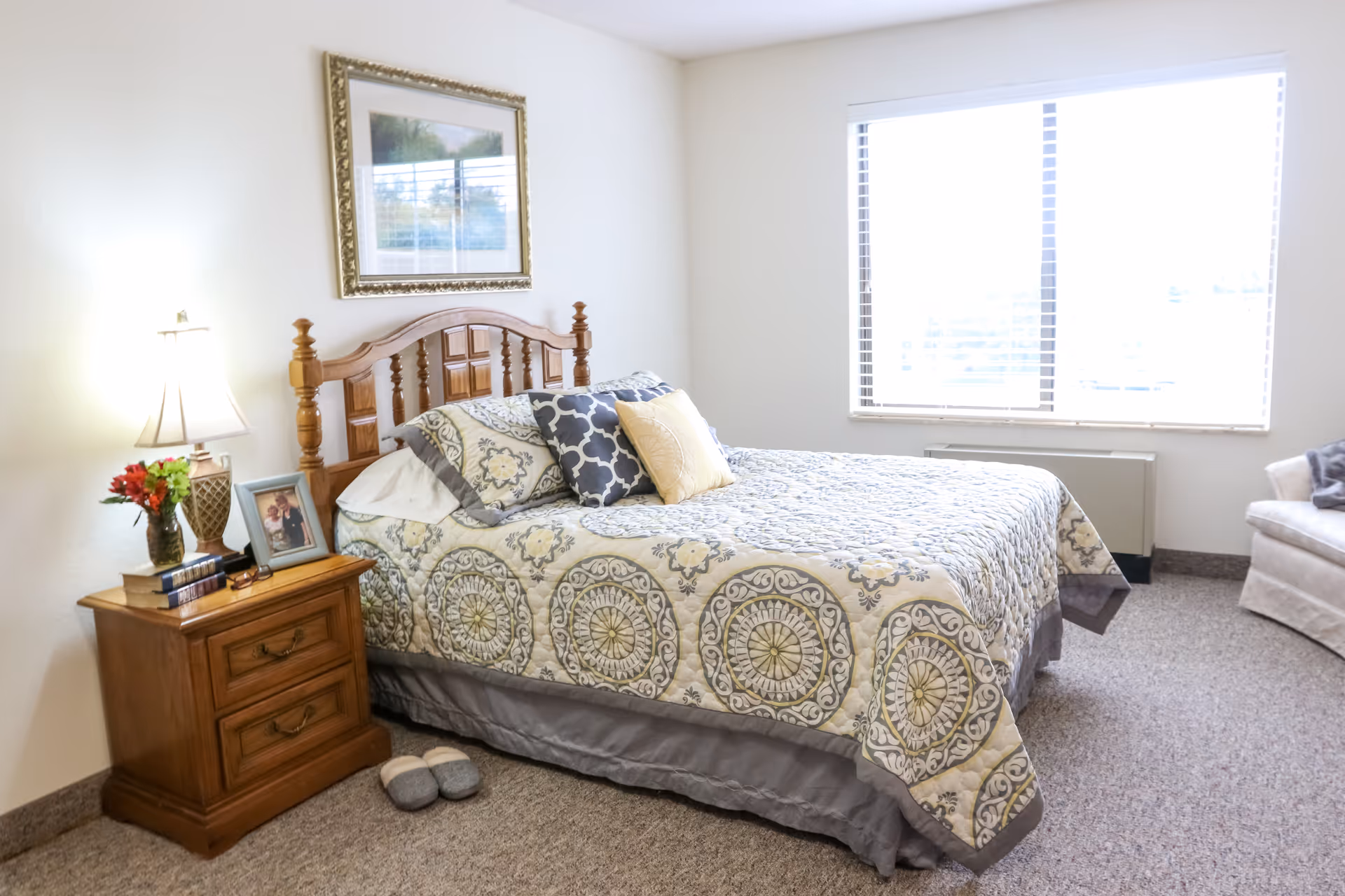 Well-lit bedroom with a patterned bed and wooden headboard, nightstand with lamp and photo, and a large window.
