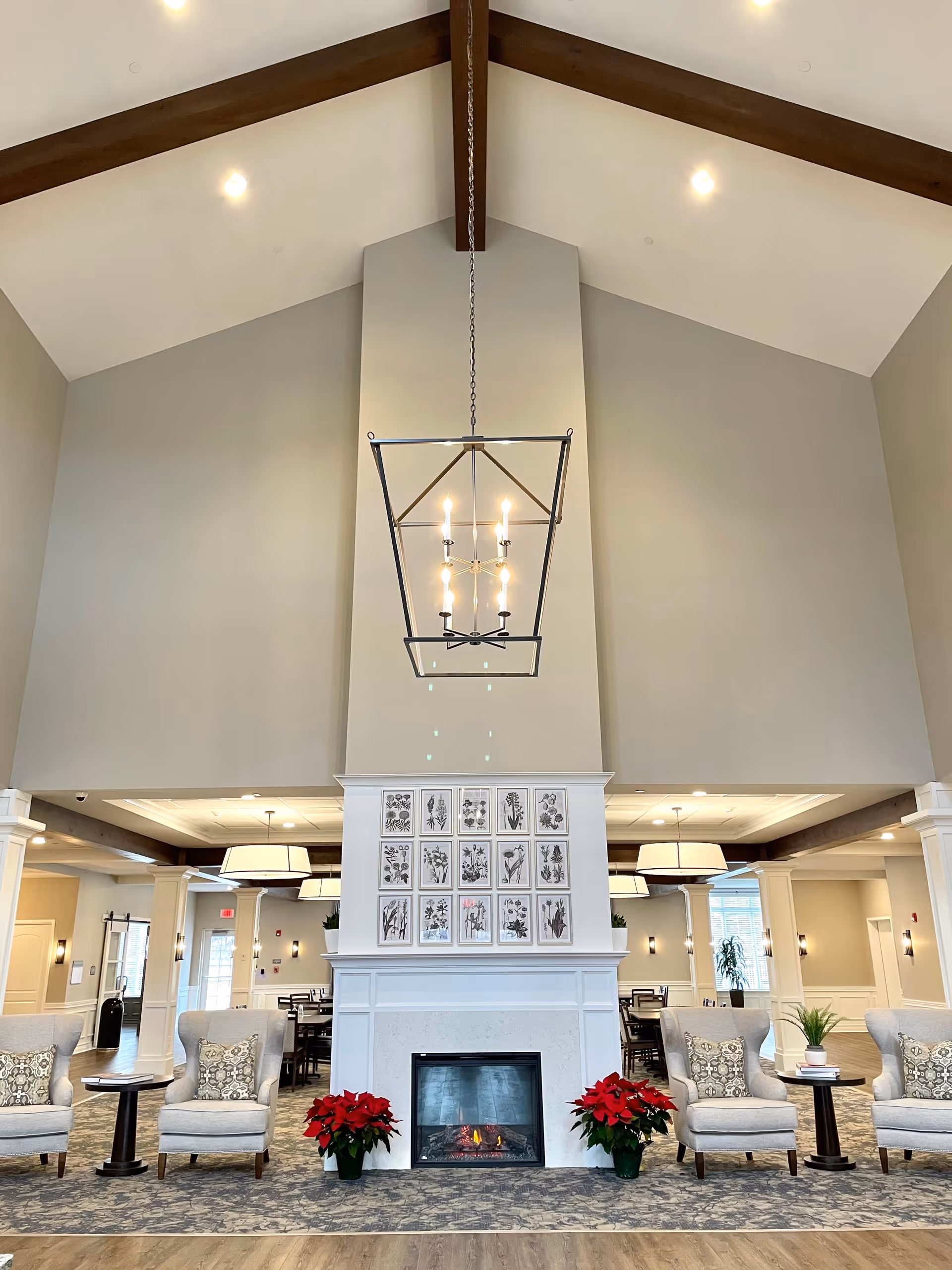 Spacious community lounge with vaulted beamed ceiling, hanging chandelier above a central fireplace and seating area with armchairs and poinsettias.