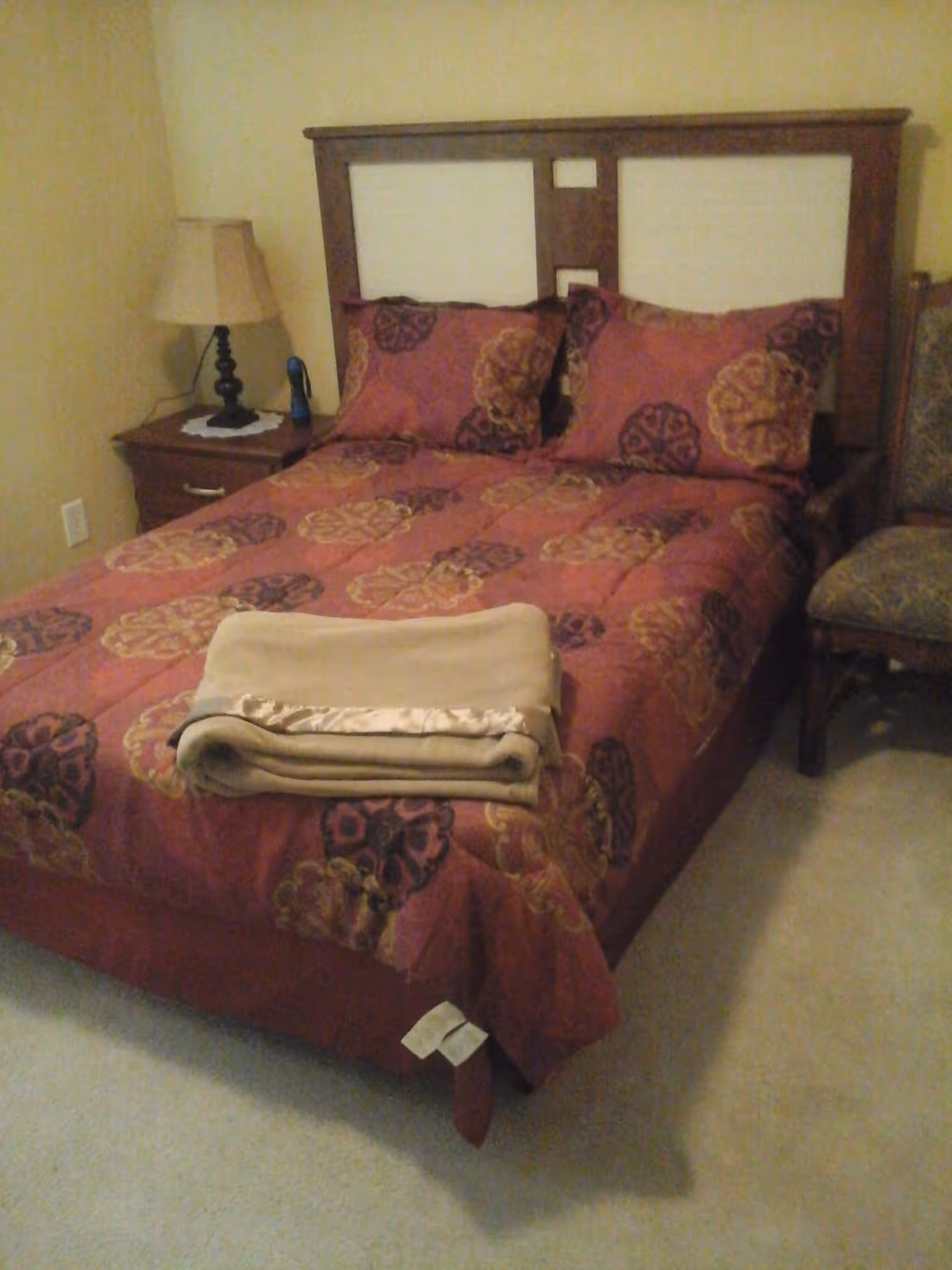 A bedroom with a bed featuring a wooden headboard and a red and gold patterned bedspread with matching pillows. A folded beige blanket is placed at the foot of the bed. To the left of the bed is a wooden nightstand with a lamp and a small blue object. To the right of the bed is a cushioned chair with a patterned fabric. The walls are painted light yellow and the floor is carpeted in a light color.