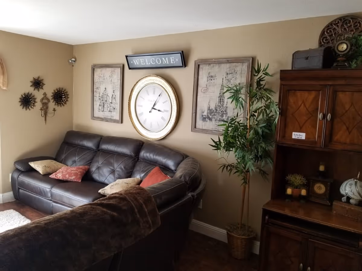 A cozy living room with a dark brown leather sectional sofa adorned with several throw pillows. Above the sofa, there is a large round clock with a gold frame flanked by two framed architectural prints and a rectangular 'WELCOME' sign. To the right, there is a tall potted plant next to a wooden cabinet decorated with small plants, candles, and a vintage-style clock.