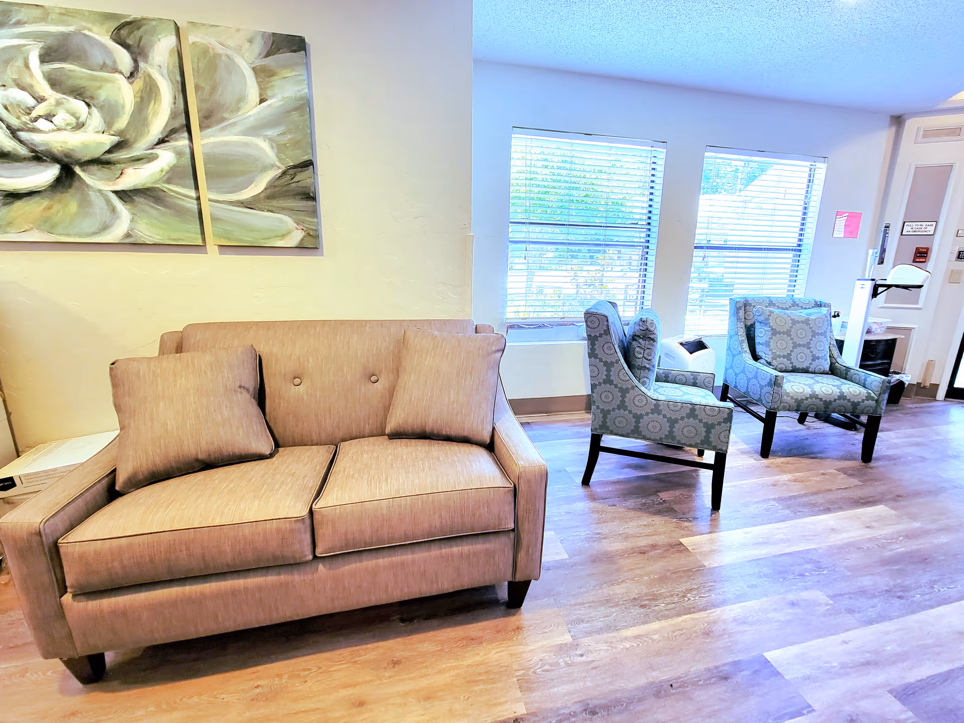 A cozy seating area in a wellness and rehabilitation facility featuring a beige loveseat with two matching pillows, two patterned armchairs, and large windows with blinds letting in natural light. The room has wooden flooring and light-colored walls adorned with a two-panel floral painting.