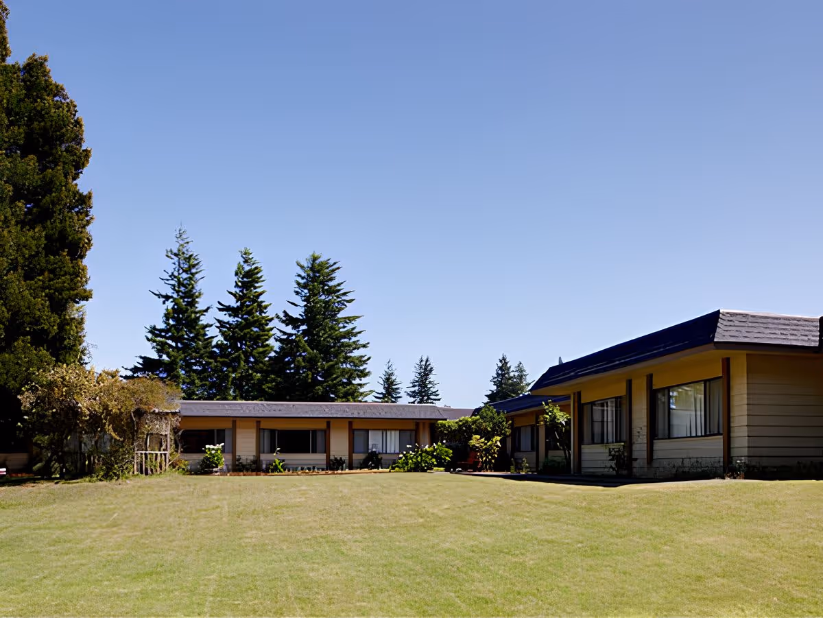 Single-story building with a wide grassy lawn, shrubs, and tall evergreen trees under a clear blue sky.