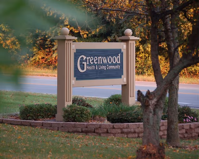 A wooden sign for Greenwood Health & Living Community situated outdoors with trees and bushes around it, and a road in the background.