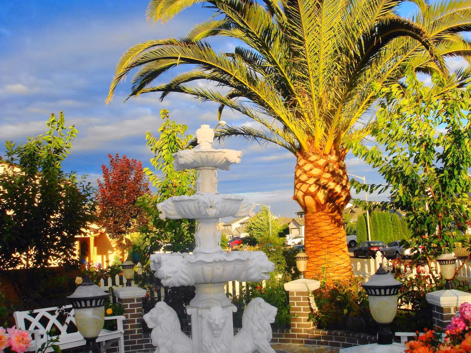 Ornamental white tiered fountain in a landscaped outdoor courtyard with a large palm tree, benches, lamps and surrounding plants.